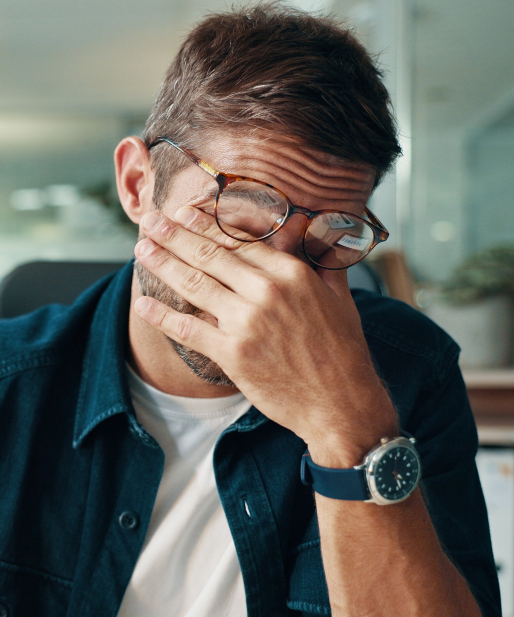 Mann mit Brille sitzt am Tisch, stützt die Hand ins Gesicht und wirkt nachdenklich oder erschöpft; trägt Hemd, T-Shirt und Armbanduhr.